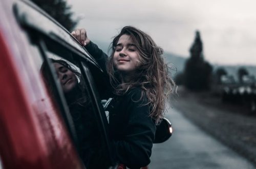 Contented young female with brown hair fluttering in wind leaning out of window of moving car in countryside