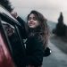 Contented young female with brown hair fluttering in wind leaning out of window of moving car in countryside